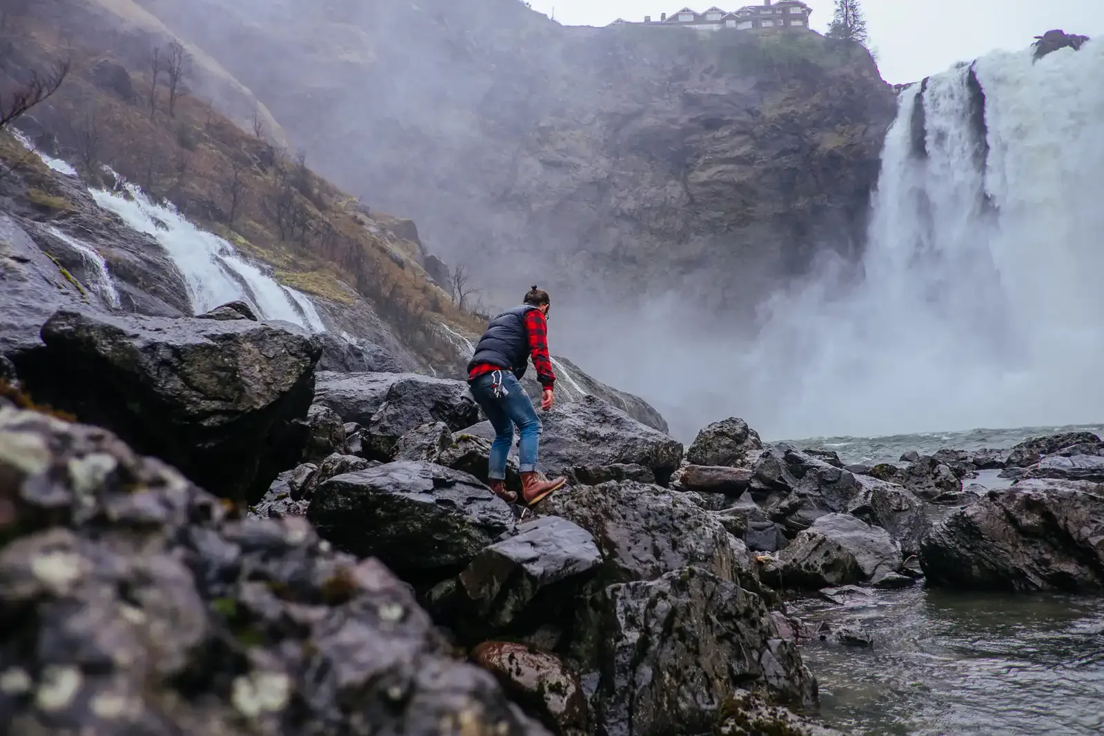 A hiker climbing over wet boulders toward a large waterfall in the mist